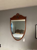 Framed oval wall mirror with wooden carved crest reflecting bed and window in room.