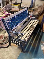 Full view of patio bench showing metal frame, blue lattice backrest design, peeling white wooden slats for seat and backrest, set indoors on concrete floor.