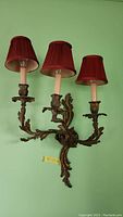 Front view of one ornate metal sconce with three battery-operated candle lights with red fabric shades mounted on a green wall.