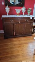 Front view of dark wood server showing two top drawers with metal handles, and three bottom cabinet doors, one with a tassel key decoration. Top surface shows signs of wear. Server is placed against red wall with vases on top (not included).