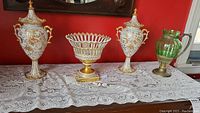Pair of two white gilt-decorated porcelain urns, pedestal bowl and green glass pitcher side by side on lace cloth