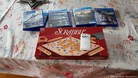 Seven PlayStation 4 video game cases laid out on a floral-patterned cloth, with a Scrabble board game box in front.
