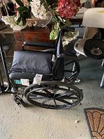 Black Hudson Home and Health wheelchair shown folded with large rear wheels and padded armrests, surrounded by flowers and other items in garage