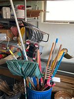 Various yard and gardening tools grouped in a bucket showing a green leaf-shaped broom, metal rake, red-handled tool, and other assorted tools.