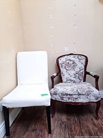 Photo shows two chairs side by side, one white upholstered with simple wood legs, the other a floral upholstered armchair with carved wood frame and arms.
