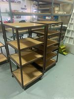 Front perspective view of two commercial shelving/display units with metal frames and wooden shelves on caster wheels, placed on a smooth floor in a warehouse setting.