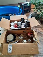 Two boxes with various ceramic mugs, glasses, and plates. First box contains mainly ceramic mugs, second box has glasses, jars, plates.
