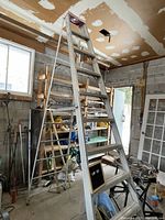 Tall aluminum A-frame step ladder set up inside a garage with shelving and other tools around it. Visible wear and rubber feet on the legs.