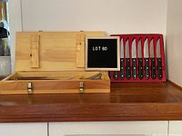 Wooden BBQ case open displaying slots for 4 BBQ tools; unopened Henkel's steak knife set in original packaging beside it.