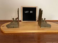 Photo of the pair of brass bookends mounted on a wooden plank, showing overall front and side profile and the 'LOT 93' sign between them.