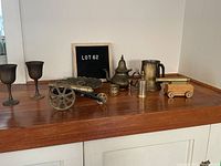 Full view of the lot including brass goblets, miniature cannon replica, teapot, pitcher, and other decorative brass items on wooden surface.
