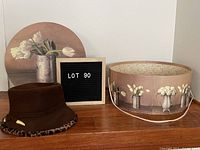 Dark brown felt winter hat with leopard print lining and gold-tone decorative pin shown next to matching floral hat box and a wooden letter board displaying 'LOT 90'.
