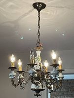 Front view of the chandelier showing six lit candle style bulbs, ceramic floral motifs on the arms and central column, and dark metal scrollwork.