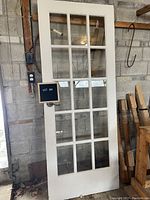Front view of the white painted French door leaning against a concrete wall showing its 15 glass panes, metal handle, and hinges.