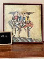 Frontal photo showing the framed art with three women riding unicycles, with a visible artist signature and red-brown frame