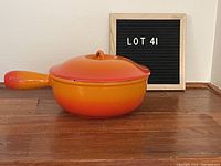 Side view of the Le Creuset fondue pot with lid in flame orange color on wooden surface with display board in background.