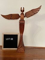Front view of hand carved wooden eagle totem with wings spread wide on a flat surface next to a sign indicating lot number.