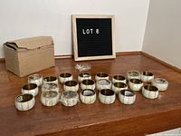 23 mother of pearl and brass napkin rings arranged on wooden surface near box and black sign reading Lot 8.