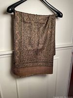 Pashmina shawl hanging on a black hanger showing detailed intricate paisley design in brown tones with pink hues, draped to reveal pattern and texture.