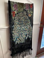 Front side showing an embroidered peacock with vibrant, detailed feathers and floral designs, velvet-like edges, and tassels.