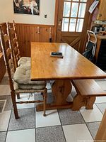 Photo showing pine dining table with matching bench and two wooden chairs with cushions, set in a kitchen area with tiled floor and wood paneling.