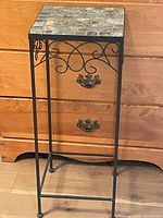 Full view of black metal plant stand with faux stone top, showing scrollwork on sides and overall shape.