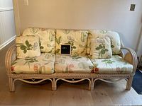 Front view of light-colored rattan sofa with floral cushions and throw pillows, showing detailed braided rattan frame and cushion pattern.