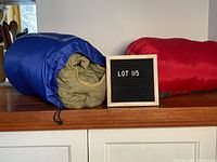 Two sleeping bags side by side on a wooden surface; one green/khaki inside a blue storage sack, the other red inside a red storage sack.