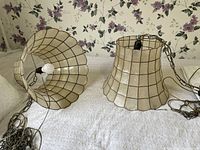 Photo showing two swag lamp shades laid on a white textured surface with floral wallpaper background. Both shades have a translucent, cream-colored material with a metal grid and chains for hanging.