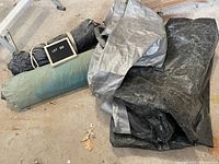 Four tarps in various states, including rolled green and black tarps, and folded gray and black tarps, on a warehouse floor.