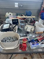 Overview of assorted used tools and items on table including chains in plastic bin, red bottle jack, floodlights, and container with small handheld tools and hardware.