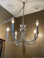Five-arm white metal chandelier with candle-style bulbs lit, showing decorative curled metal arms and chain suspension against brown wall and ceiling.