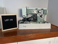 Front and side view of vintage Viking sewing machine Model 714 in green, showing controls and power cord, seated on cream base with original case under the machine.