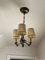 Front view of the three-bulb candle-style light fixture with beige fabric shades and brass-colored metal frame hanging from the ceiling chain. Shades show wear.