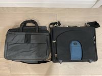 Two laptop bags side by side on a wood floor; one gray with multiple compartments and one black with blue front panel and shoulder strap.