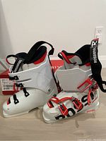 Pair of white Rossignol Hero Racing Team ski boots with red and black accents, buckles, straps, and padding visible, placed on floor next to red Rossignol box.