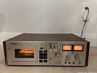 Front view showing cassette compartment with cassette inside, buttons, knobs and meters on silver face with wood side panels.