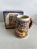 Front and side view of ceramic 1992 Oktoberfest Budweiser beer stein with colorful raised relief and pretzel-shaped handle, with original box in background.