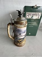 Front view of the ceramic beer stein showing painted scene of peregrine falcons with pewter lid and handle, beside original green box.