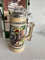 Front and side views of a new ceramic beer stein with detailed train-themed artwork, ornate handle, and pewter lid shaped like a steam locomotive, with original box.