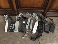 Overview of all power strips and surge protectors in the lot arranged on carpet in front of wooden furniture. Various colors and outlet counts visible.