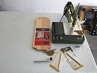 Overview photo of the full lot showing the Proxxon Micromot Deluxe scroll saw, natural pine plaque, angle guide, saw blades, and other accessories on a table.