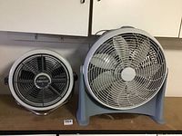 Side-by-side image of two small fans on a surface under a cabinet, one smaller Seabreeze Turbo Aire and one larger Lakewood fan