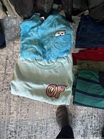 Photo of assorted men's large clothing items including t-shirts, tops, and sweaters folded on the floor, showing details of prints and colors.