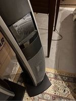 Front angled view of tower heater showing mesh grille and power cord, positioned next to table and boxes.