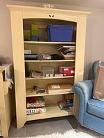 Front view of wooden bookshelf in hay color with four shelves containing various items not included in the lot. Shows bookshelf in a room next to a blue upholstered chair.