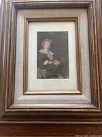Framed painting showing a young boy with curly blond hair dressed in green historical costume, sitting and holding an object, surrounded by a wooden frame and beige mat.