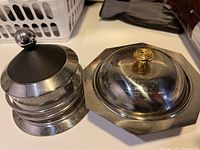 Two stainless steel parmesan cheese bowls with lids. One round with black knob lid, the other octagonal with gold knob lid.