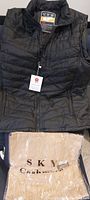Black heated vest laying flat with tan cashmere scarf packaged in clear plastic at base.