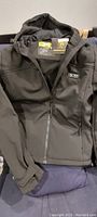 Black hooded heated jacket folded on fabric surface with visible front zipper and thumbhole adjustable sleeve cuffs.